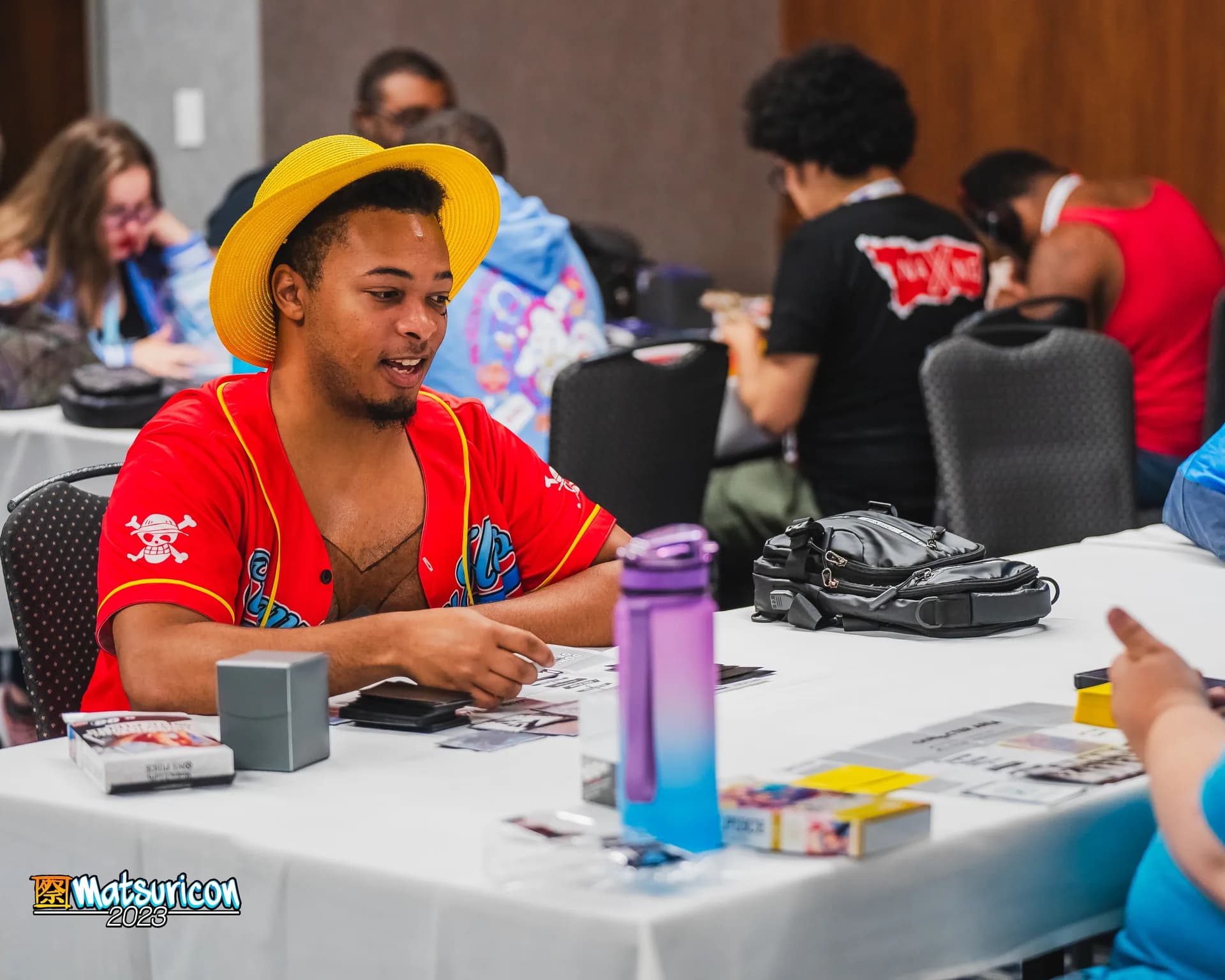 A smiling Matsuricon attendee in a straw hat plays cards at a crowded table.