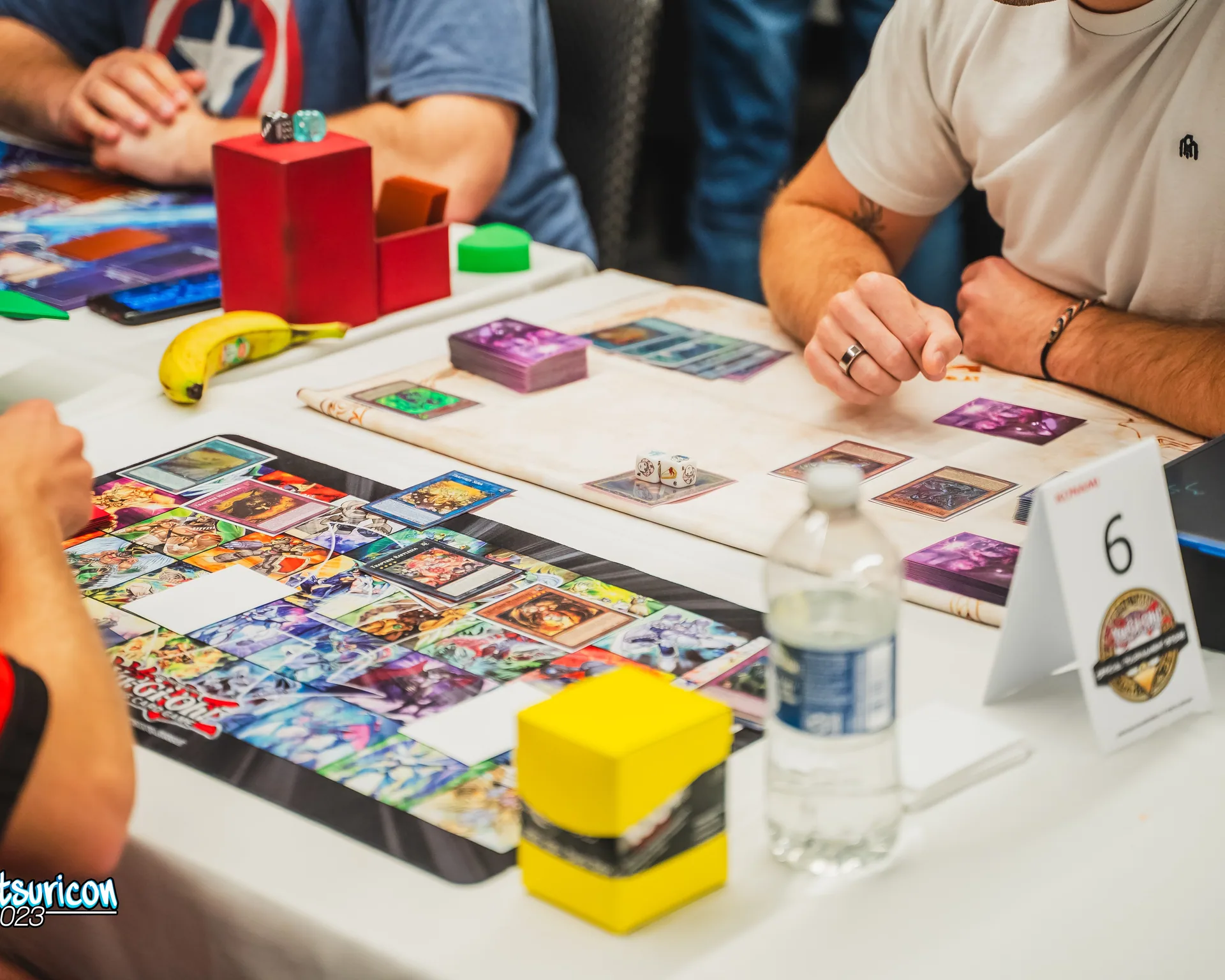 Trading card game competitors face each other across a table covered with decks, cards, dice, and colorful playmats.