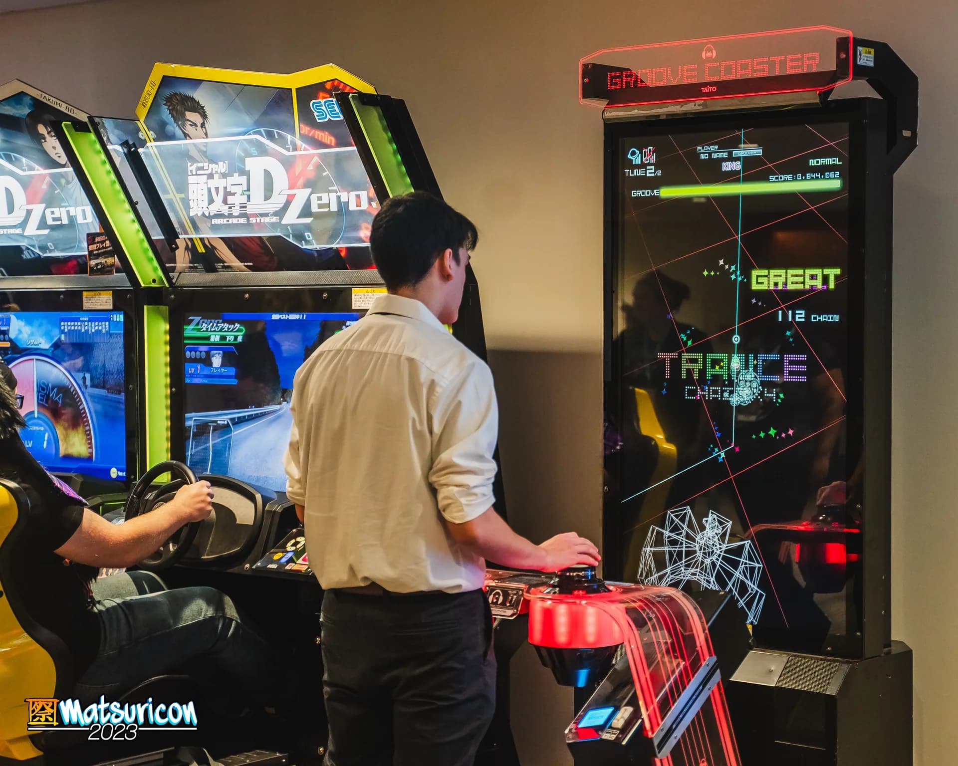 Two Matsuricon attendees play brightly lit Japanese racing and rhythm arcade cabinets.