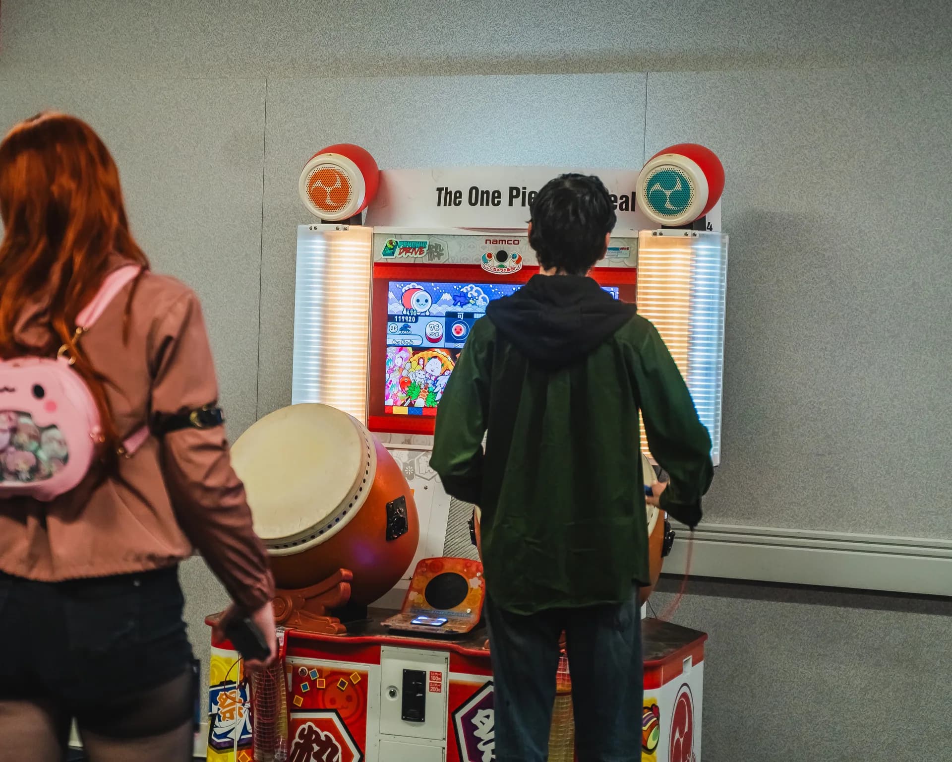 A Matsuricon attendee plays a Taiko no Tatsujin drum rhythm arcade machine while another attendee watches.
